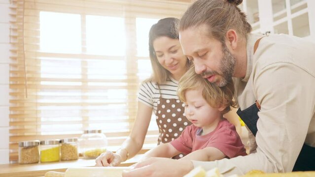 Caucasian father and mother teaching baby son kneading dough on kitchen counter at home. Parents and child boy kid enjoy and fun indoors activity cooking pizza together. Family relationship concept.