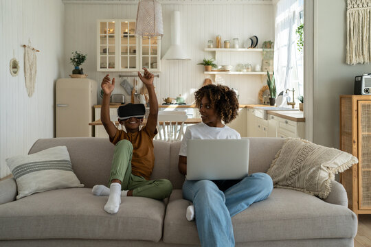 African American family mom and son sit on sofa with electronic devices. Mother-parent sits with laptop, son in virtual reality glasses. Black boy waves hands in gaming helmet. Family and technology - Powered by Adobe
