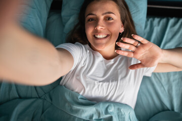 Beautiful young woman taking selfies at home in bed. Blogger influencer having a facetime video call.