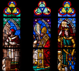 St. Denis Basilica Paris, France. Set of three colorful and detailed medieval era stained glass panels depicting saints set in stone gothic tracery in a cathedral. Illuminated windows against a black