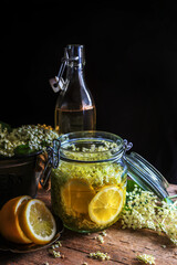 Homemade elderflower syrup in jar and elderberry flowers on wooden table - dark and moody photography