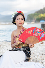 Fototapeta premium Portrait of a cute girl in a beautiful off-the-shoulder costume and a crown in her hair posing by the seashore with red hand fan