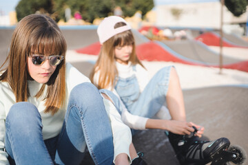 Two teenage blonde sisters are sitting in a pump track park adjusting their inline skates.