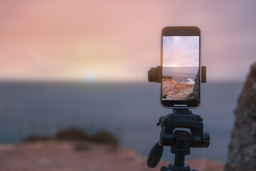 the phone on a tripod stands at dawn on a mountain by the sea and photographs the dawn. A beautiful seascape has a place for an inscription close-up on the phone - obrazy, fototapety, plakaty