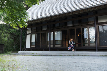 浴衣を着て日本家屋の縁側で見上げてくつろぐ、うちわを持った中年女性　広角の全身