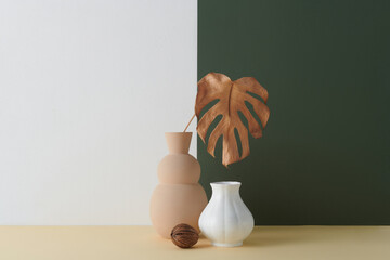 Horizontal shot of contemporary still life composition of two vases, brown dried seed and Monstera leaf against white and dark green wall background