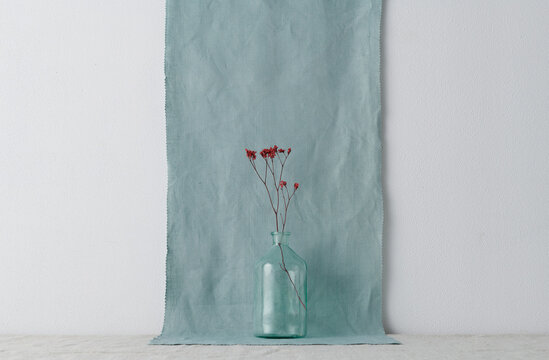 Horizontal shot of modern still life installation with glass vessel and dried red flowers against white wall and blue linen fabric background, copy space