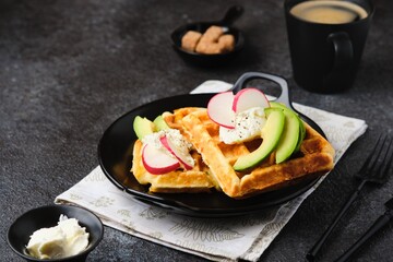 Unsweetened cheese waffles with cream cheese, avocado and radish slices on a black plate on a dark concrete background.