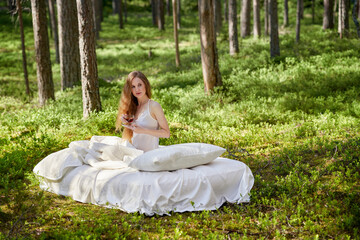 Woman wakes up in a summer forest. A girl drinks morning tea sitting on a mattress in the woods