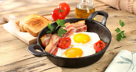 fried eggs with bacon and tomatoes in a frying pan. breakfast on the wooden table. sunny morning. country style. organic farm food