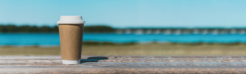 Disposable paper coffee cup on wooden bench outdoors,paper cup of coffee in the park on a wooden bench.lake in the background.copy space.Summer day.Banner,advertisement.Toned.