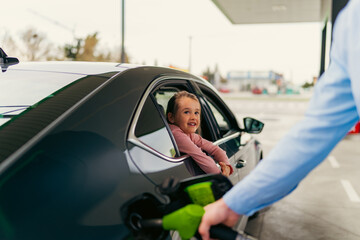 The daughter sits in the car leaning against the window and watches her father pour fuel into the tank
