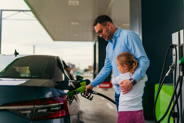 The father pours fuel into the car while his daughter hugs him and keeps him company, younger...