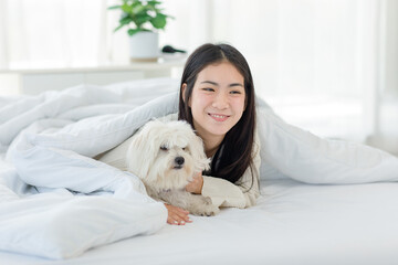 Asian young happy cheerful female owner laying lying down smiling hugging cuddling embracing best friend companion dog white long hair mutt shih tzu under blanket on bed together in bedroom at home