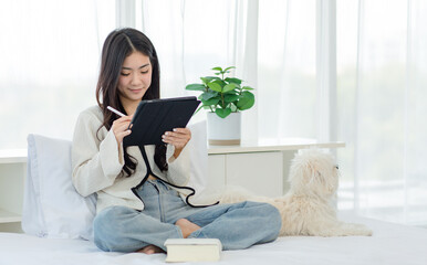 White long hair mutt shih tzu dog laying lying down on bed with Asian young cheerful female owner sitting smiling using touchscreen tablet computer on blurred background in bedroom.