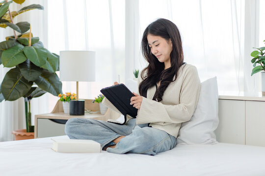 Asian young cheerful female teenage model sitting smiling on white clean sheet bed using pen with touchscreen tablet computer browsing surfing internet shopping working online in modern decor bedroom