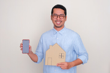 A man smiling and showing blank mobile phone screen while holding house shape cardboard - obrazy, fototapety, plakaty
