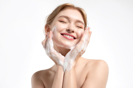 A happy young woman cleanses her skin with a gentle facial foam, the model applies the cleanser to her skin and smiles.