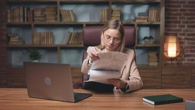 Hard-working financial director in glasses reading annual report about company income. Focused woman sitting at table with laptop in cozy office and working till late evening.