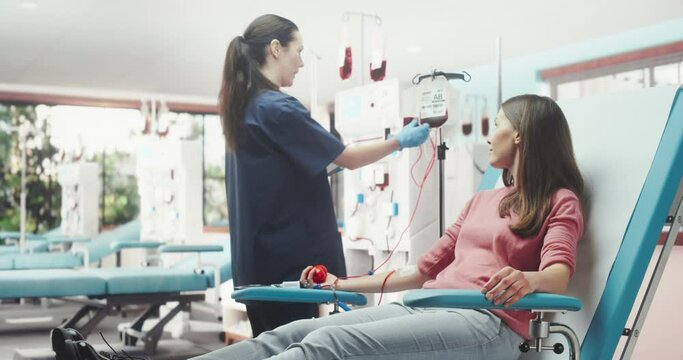 Caucasian Woman Donating Blood For People In Need In Bright Hospital. Female Nurse With Tablet Computer Coming In To Check Progress And Well-Being Of Donor. Donation For Organ Transplant Patients.