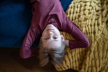 Portrait teenage boy lying on sofa upside down with hands behind head and looking camera. Smile on...