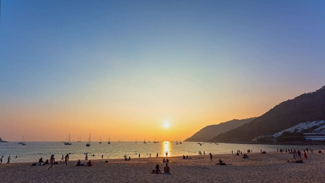.time lapse golden sky at sunset above the sea at Nai Harn beach..Yachts are on the horizon of the sea during beautiful golden sky..crowd of tourists at Nai Harn beach in wonderful sunset..