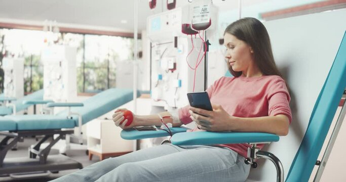 Caucasian Woman Donating Blood For People In Need In Hospital. Female Donor Squeezing Heart-Shaped Red Ball To Pump Blood And Chatting Online Using Smartphone. Donation For Victims Of Accidents.