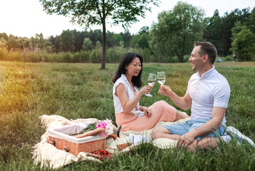 Happy diverse couple relaxing on blanket, having picnic in sunny garden, holding wine glass.