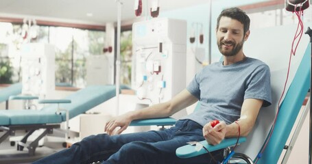 Caucasian Man Donating Blood For People In Need In Bright Hospital. Male Donor Squeezing Heart Shaped Red Ball To Pump Blood Through The Tubing Into The Bag. Donation For Burn Victims Patients.