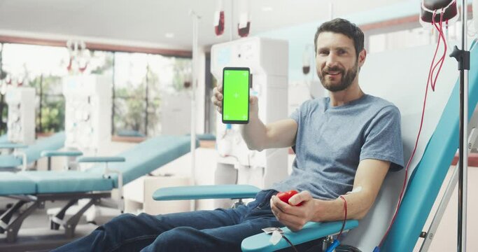 Blood Donation Center Advertisement Mock Up: Caucasian Man Sitting In The Chair, Squeezing Red Ball To Pump Blood Into Bag And Showing Green Screen Chromakey Smartphone To Camera. Become a Donor Ad.