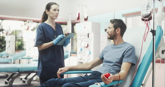 Caucasian Man Donating Blood For People In Need In Bright Hospital. Female Nurse With Tablet Computer Coming In To Check Progress And Well-Being Of Donor. Donation For Heart Surgery Patients.