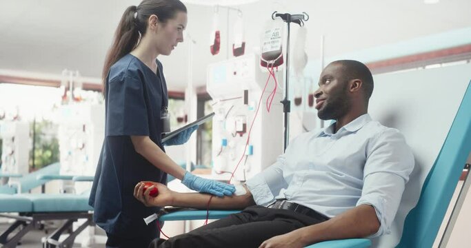 Black Businessman Donating Blood For People In Need In Bright Hospital. Female Nurse With Tablet Computer Coming In To Check Progress And Well-Being Of Donor. Donation For Heart Surgery Patients.