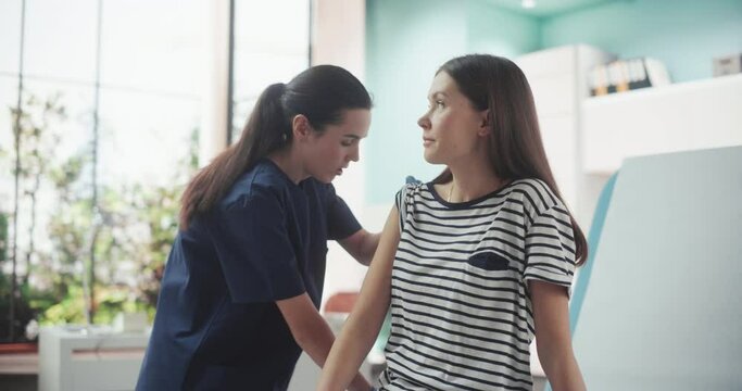 Happy Caucasian Woman Sitting In The Chair In Bright Hospital And Getting Her Flu Vaccine. Professional Female Nurse Is Performing Injection And Putting A Patch On. Public Healthcare Concept.