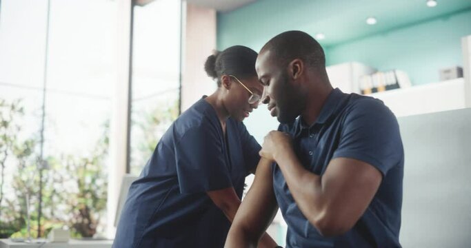 Black Man Sitting In The Chair In Hospital And Getting His Flu Vaccine. Professional African Female Nurse Is Informing Patient About Side Effects and Performing Injection. Public Healthcare Concept.