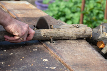 Hands of a worker sawing firewood with an industrial electric circular saw close-up. Harvesting firewood for the winter, sawmill. Sawing firewood with circular saw blade