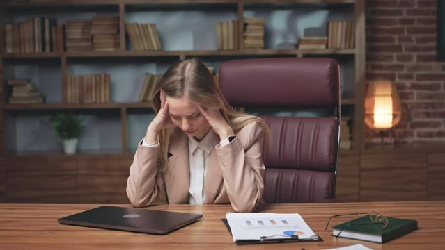 Long-haired blonde woman having strong headache and finishing work in late evening. Exhausted lady sitting at long wooden table, closing wireless gadget and holding head with hands.