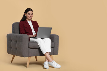 Fototapeta na wymiar Happy woman with laptop sitting in armchair on beige background, space for text