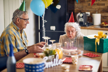 Happy senior woman blowing candles on her birthday cake, sitting at table with husband cheering her.