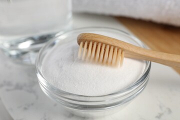 Bamboo toothbrush and glass bowl of baking soda on white marble table, closeup