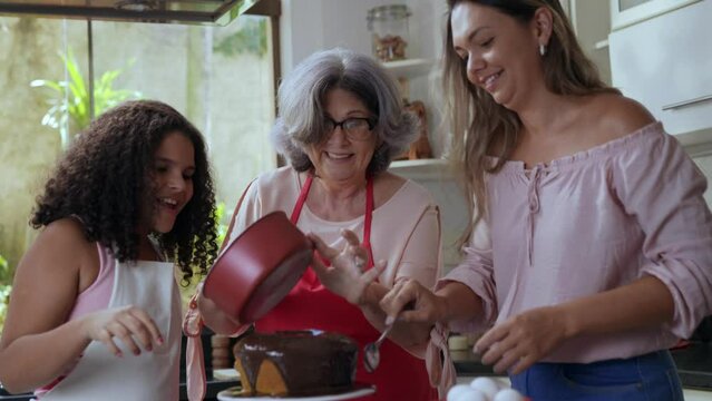 av&oacute; m&atilde;e e filha latinas na cozinha pondo cobertura no bolo que fizeram juntas para o dia das m&atilde;es