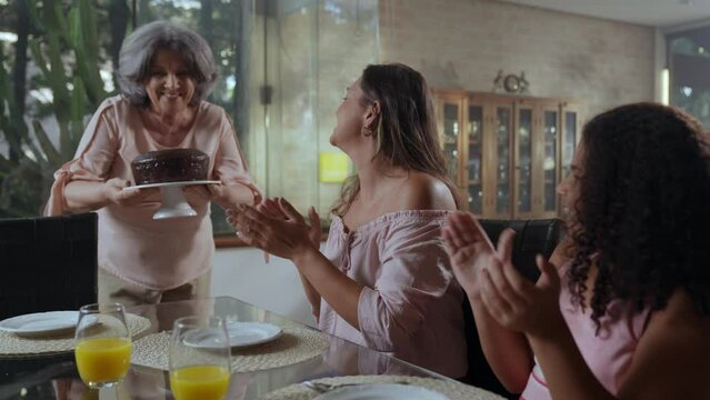 av&oacute; idosa brasileira levando um bolo at&eacute; a mesa de jantar enquanto sua filha e neta batem palmas no dia das m&atilde;es