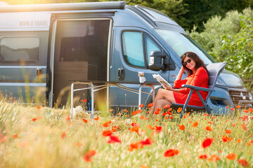 Work and Travel concept, outdoor leisure activity and wanderlust lifestyle - woman dressed in red dress reading a book in a meadow among blooming red poppies next to an off-road camper van.