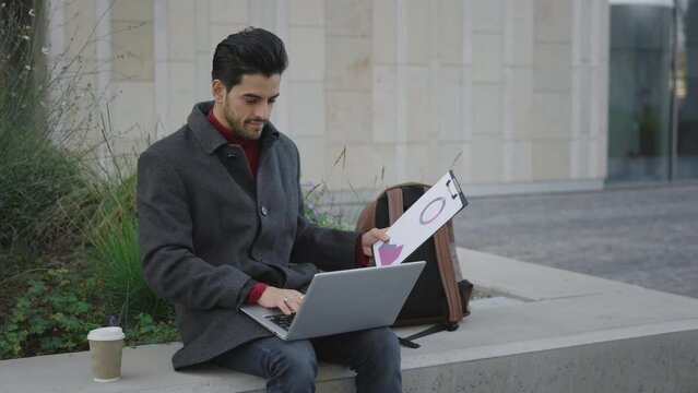 Handsome Bearded Multiethnic Man Sitting Working On Laptop Outside. Foreigh Student Studying In the Street Using Computer And Taking Notes to his Tablet