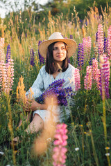 Fototapeta na wymiar Portrait of young attractive woman in dress and straw hat holding bouquet of blooming lupin flowers on summer meadow at sunset