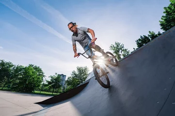 Fotobehang Low angle view of a mature tattooed man riding a bmx bike on a ramp in a skate park. © Zamrznuti tonovi