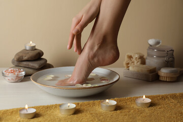 Woman soaking her foot in bowl with water and petals on white wooden floor, closeup. Pedicure procedure