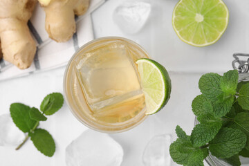Glass of tasty ginger ale with ice cube and ingredients on white tiled table, flat lay