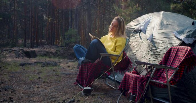 Nature as a path to mental health. Zoom in of a young woman reading a book near a tent in a pine forest. Solitude with your thoughts and nature to the singing of birds and the buzzing of insects.