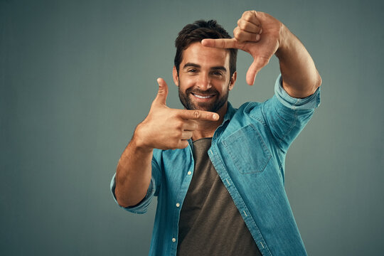 Frame, hands and face portrait of man in studio on gray background with happiness, confident and smile. Photographer, happy and male person with hand sign for picture, photography and finger border