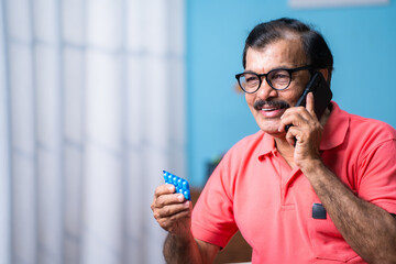 Indian senior man talking on mobile phone by holding tablets in hand at home - concept of telemedicine, elderly health care and pharmacy